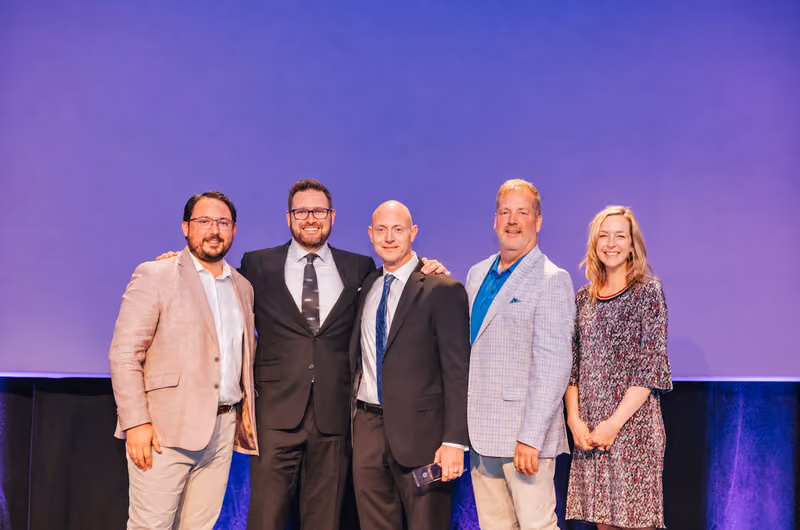 Five colleagues in business attire standing together and smiling in front of a purple stage backdrop.